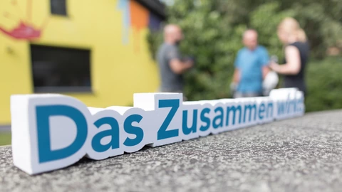Im Vordergrund steht der Schriftzug "Das Zusammen wirkt." Im Hintergrund unterhalten sich drei Personen.