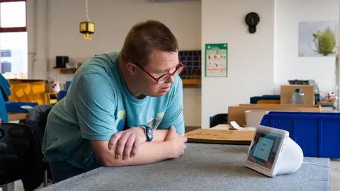 Ein Mensch lehnt mit verschränkten Armen an einem Tisch und schaut auf ein Alexa Gerät.