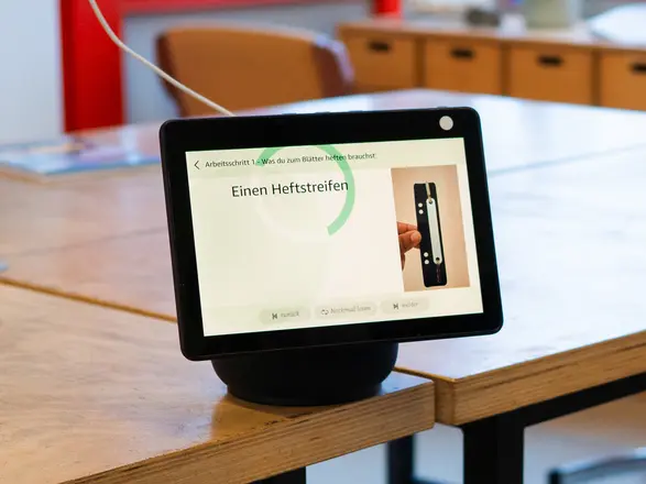 Auf dem Bildschirm eines Alexa-Gerätes ist eine Arbeitsanweisung zu sehen. Darauf steht "Einen Heftstreifen". Daneben ist ein Heftstreifen abgebildet.