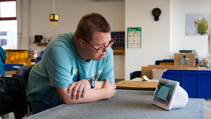 Ein Mensch lehnt mit verschränkten Armen an einem Tisch und schaut auf ein Alexa Gerät.