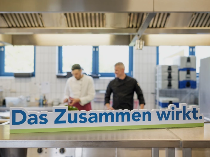 Zwei Männer in der Küche. Im Vordergrund steht der Schriftzug "Das Zusammen wirkt."